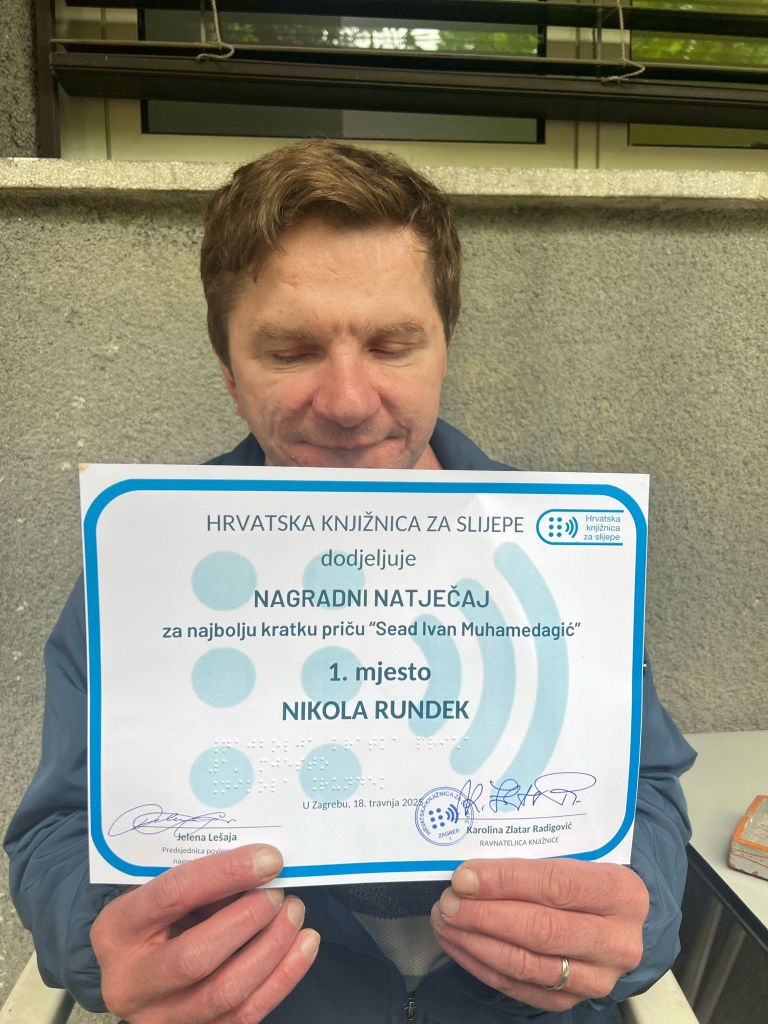 Autor priče sjedi i drži ispred sebe priznanje za osvojeno prvo mjesto natječaja Sead Ivan Muhamedagić. Na brajici piše: Najbolja priča, 1. mjesto, Nikola Rundek